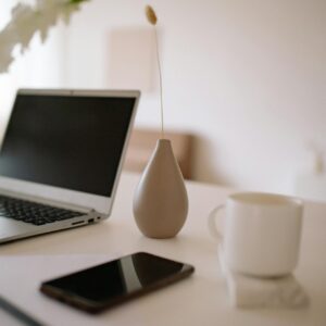 A clean and modern workspace featuring a laptop, vase, cup, smartphone, and paper on a white table.