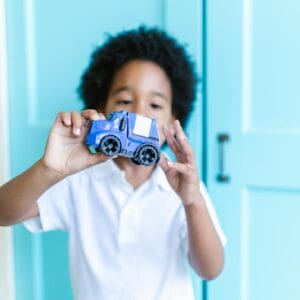 Child with afro hair playing with a blue toy truck in a bright room.