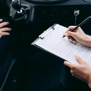 Close-up of a person writing on a clipboard inside a car, showing hands and a gear shift.
