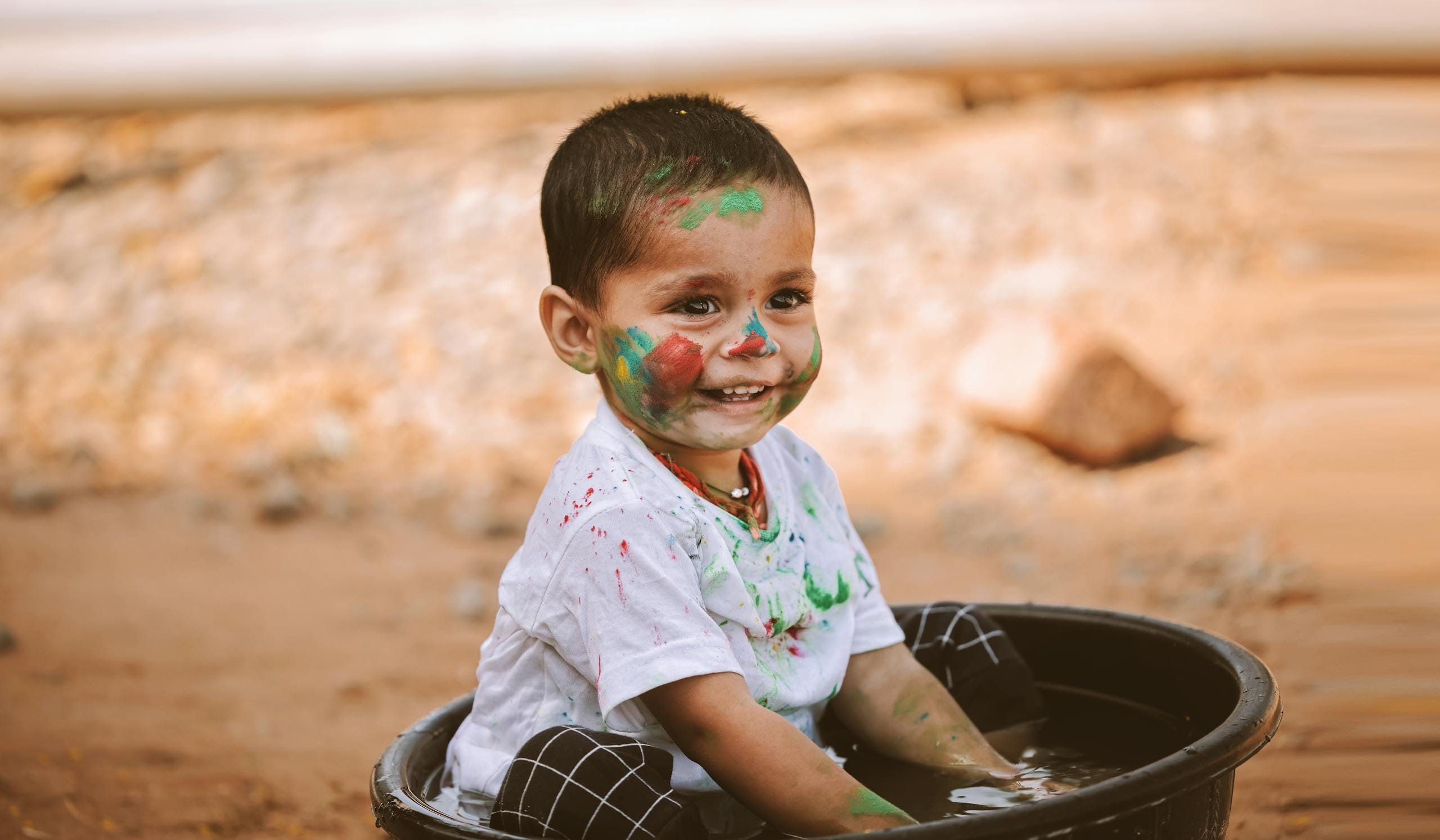 Happy child with colorful face paint playing in a basin outdoors, smiling with joyful expression.