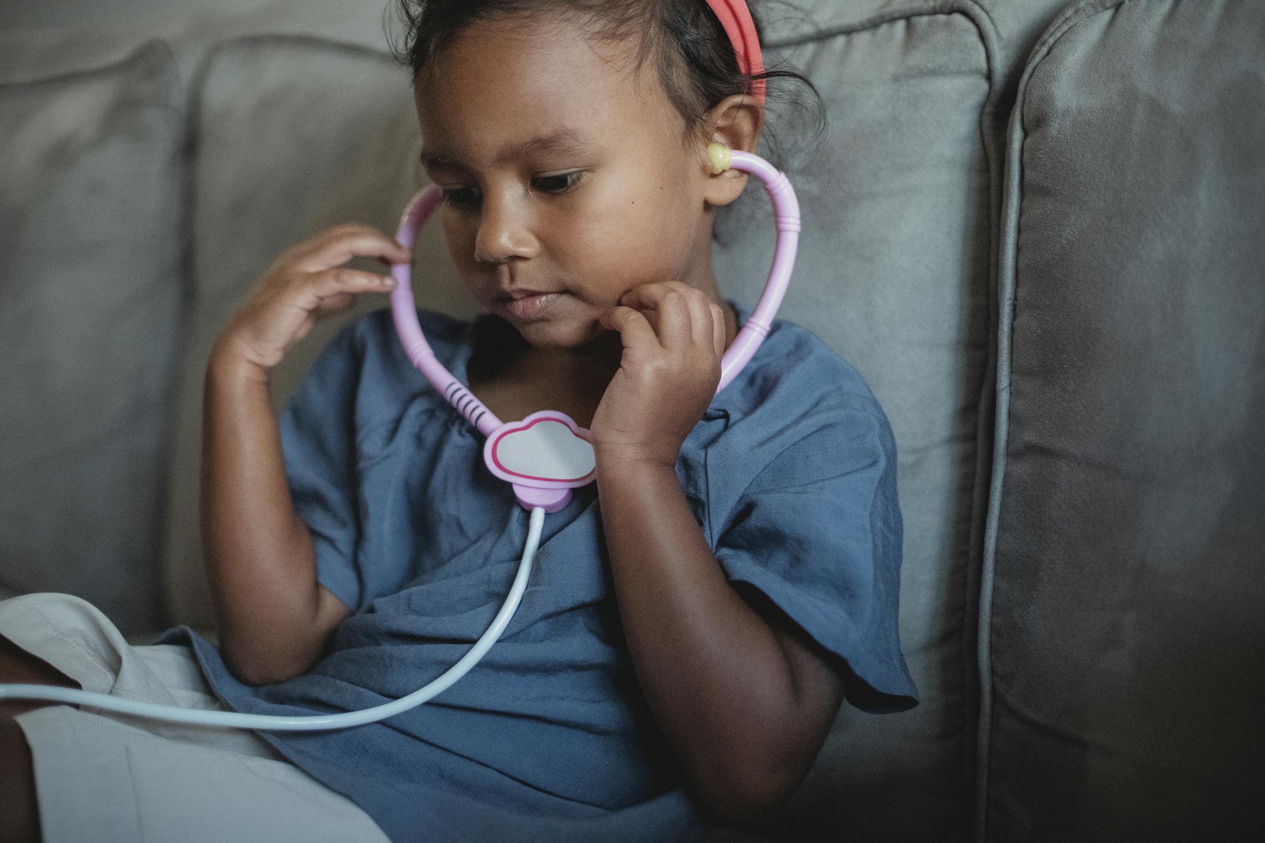 Side view of crop focused Asian kid wearing headband sitting on soft couch with toy stethoscope while playing doctor in cozy living room